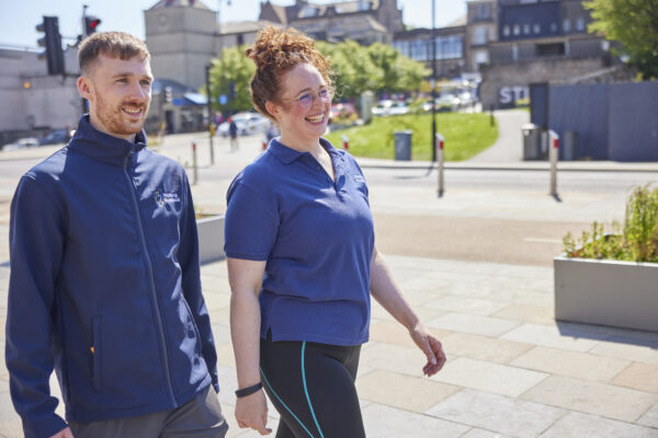 A woman and a man wearing Walking Scotland branded clothing walk side by side, smiling.