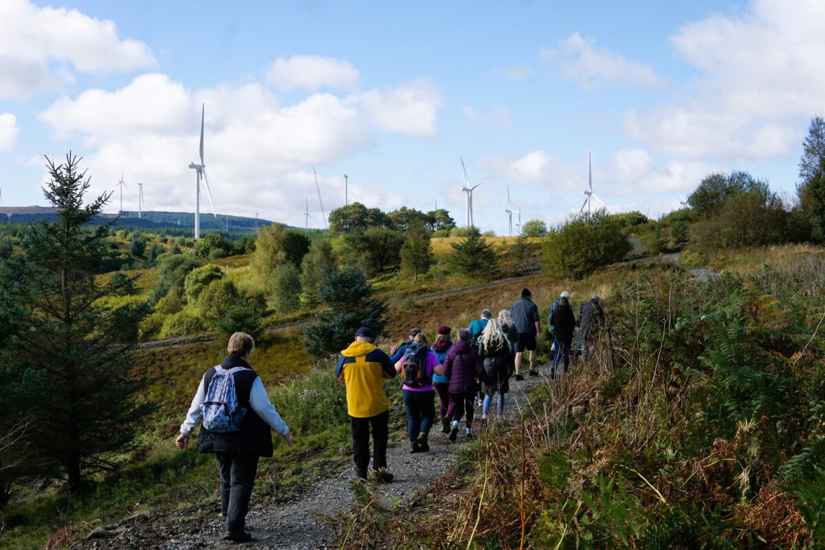 A line of people walk along a path along a gentle hillside. Wind turbines and woodland can be seen in the distance.