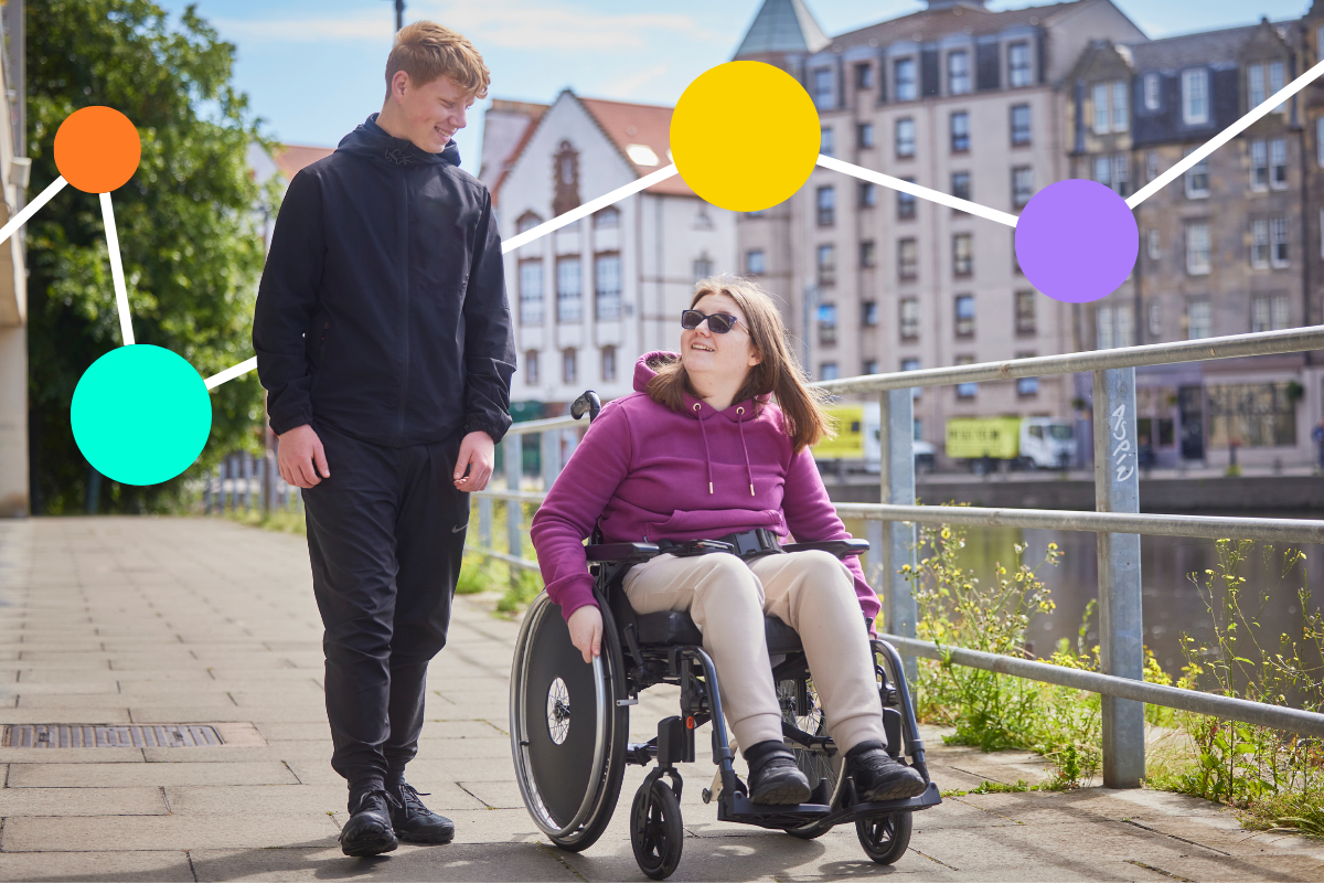 A young man smiles as he walks by a young women in a wheelchair as they walk and wheel along a riverside path. There are buildings in the background.