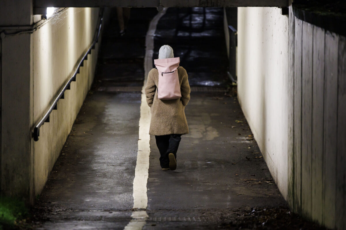 A woman walks through a lit underpass wearing a long brown coat, grey hat and pink backpack. It is nighttime and the underpass is lit up.