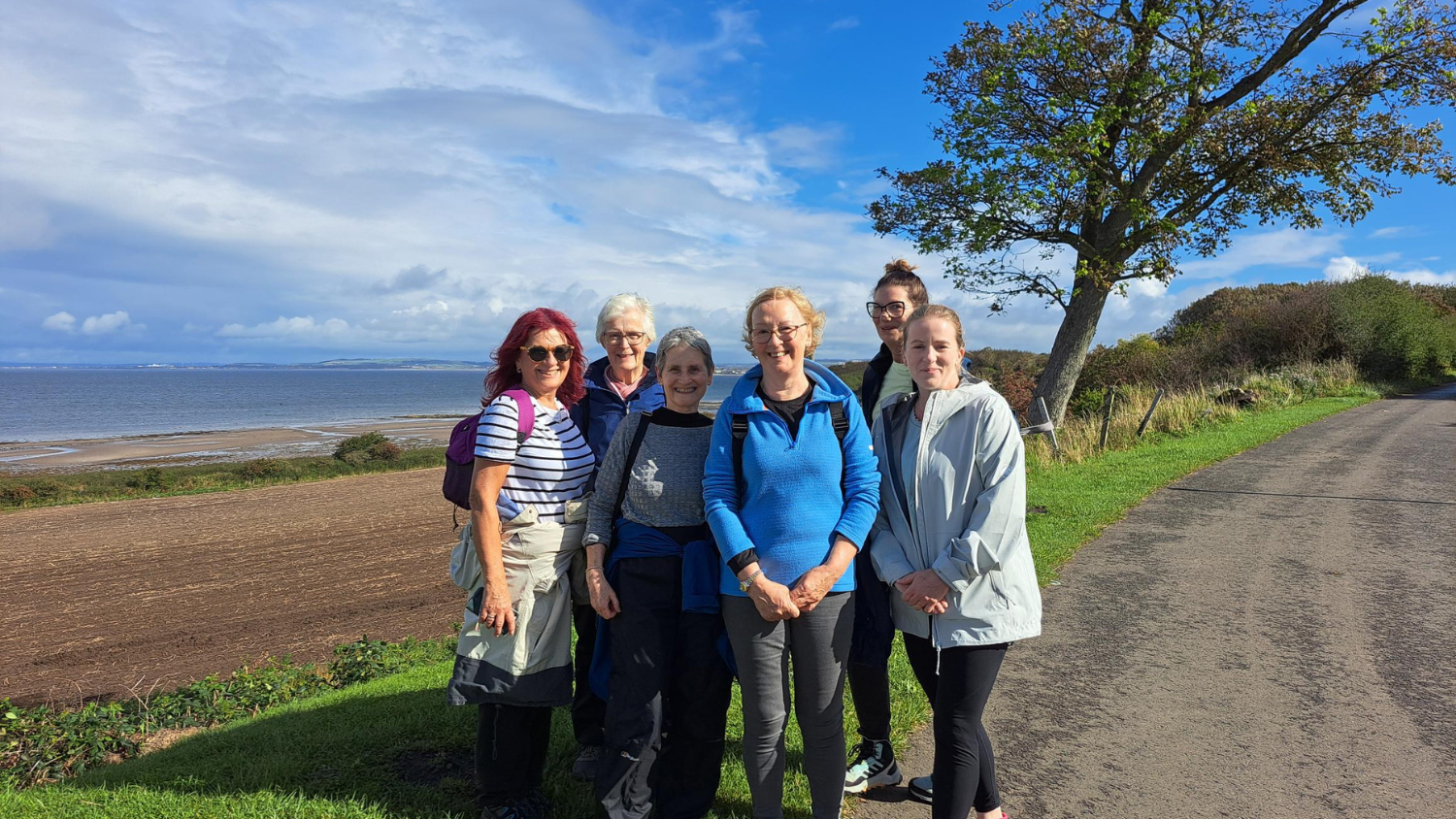 Six woman pose, smiling at the camera. There are stood to the side of a paved track. Fields and the sea are in the background. 