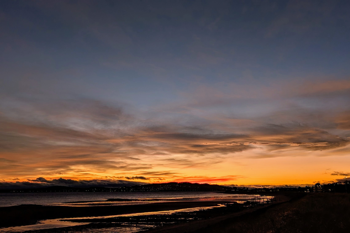 A vivid sunset over a coastal path with the lights of a town and village in the distance.