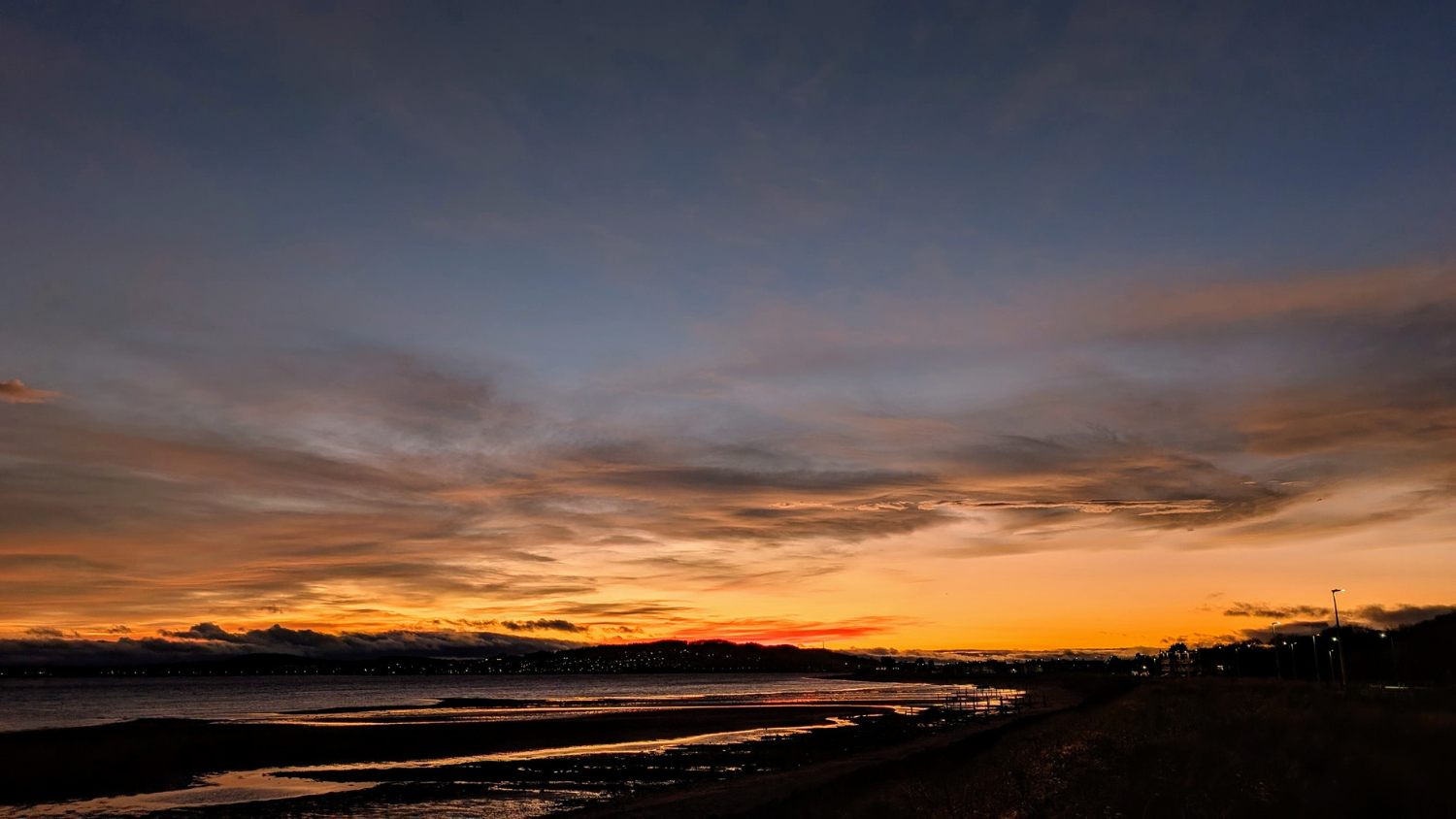 A vivid sunset over a coastal path with the lights of a town and village in the distance.