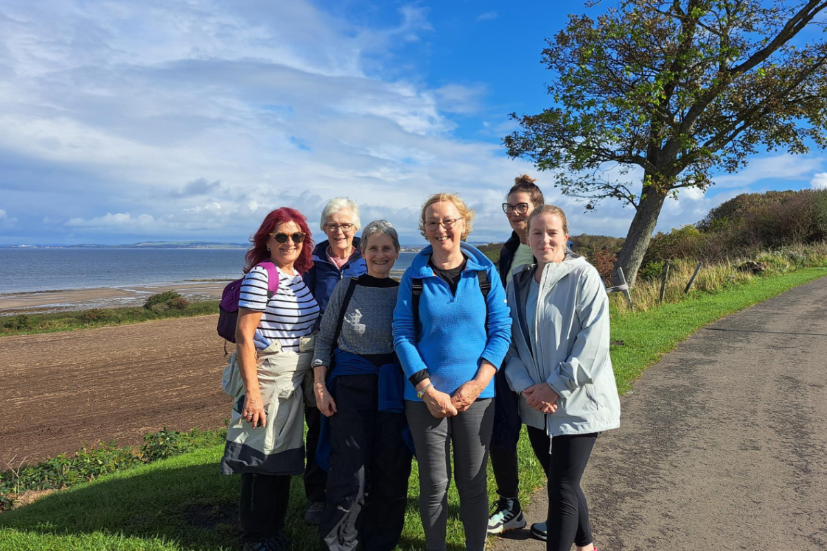 A group of women smile as they stand together on a quiet road next to a beach.