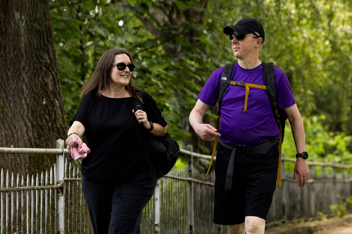 A man and woman talk as they enjoy a brisk walk along a path in a wooded area.