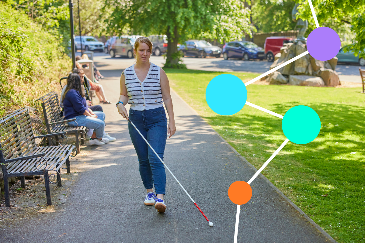 A woman with sight loss, holding a walking aid, walks along a path through an urban park. Sun shines through the nearby trees.