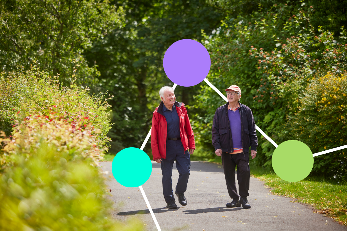 Two older gentleman smile as they walk along a woodland path, bordered by lush bushes. The sun is shining.