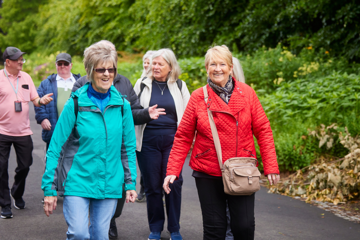 A large group of people in waterproof jackets and backpacks smile as they walk along a path bordered by trees and shrubbery,