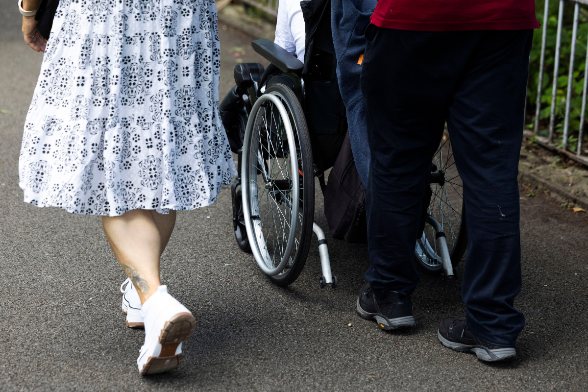 The lower half of a woman and man, walking behind an individual in a wheelchair.