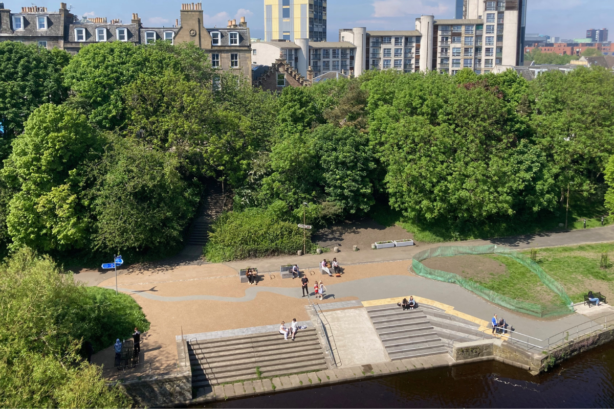 A wide-angle view over part of Coalie Park, with people walking, sitting or playing. Trees and buildings stretch into the distance.