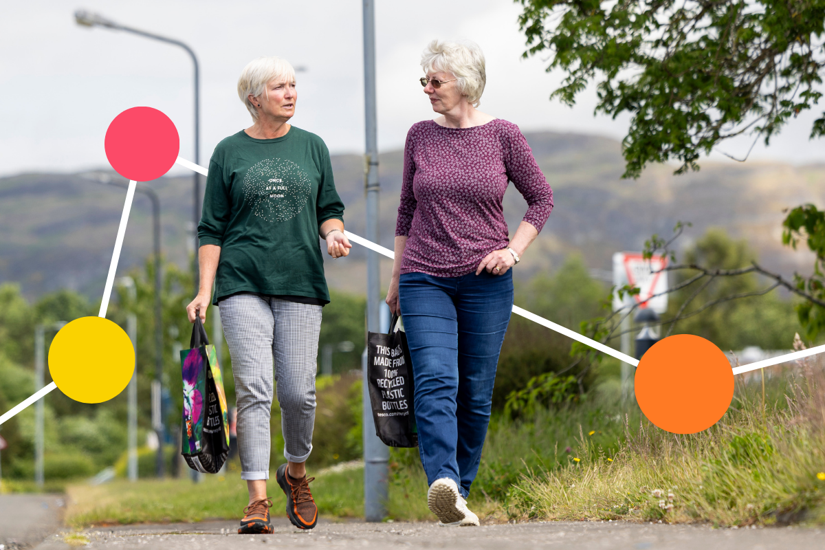 Two woman wearing light clothing and carrying bags talk as they stroll along a path.