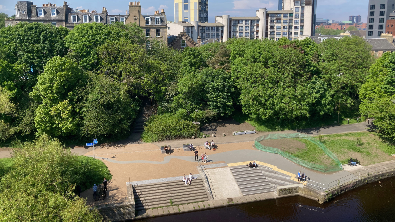 A wide-angle view over part of Coalie Park, with people walking, sitting or playing. Trees and buildings stretch into the distance.