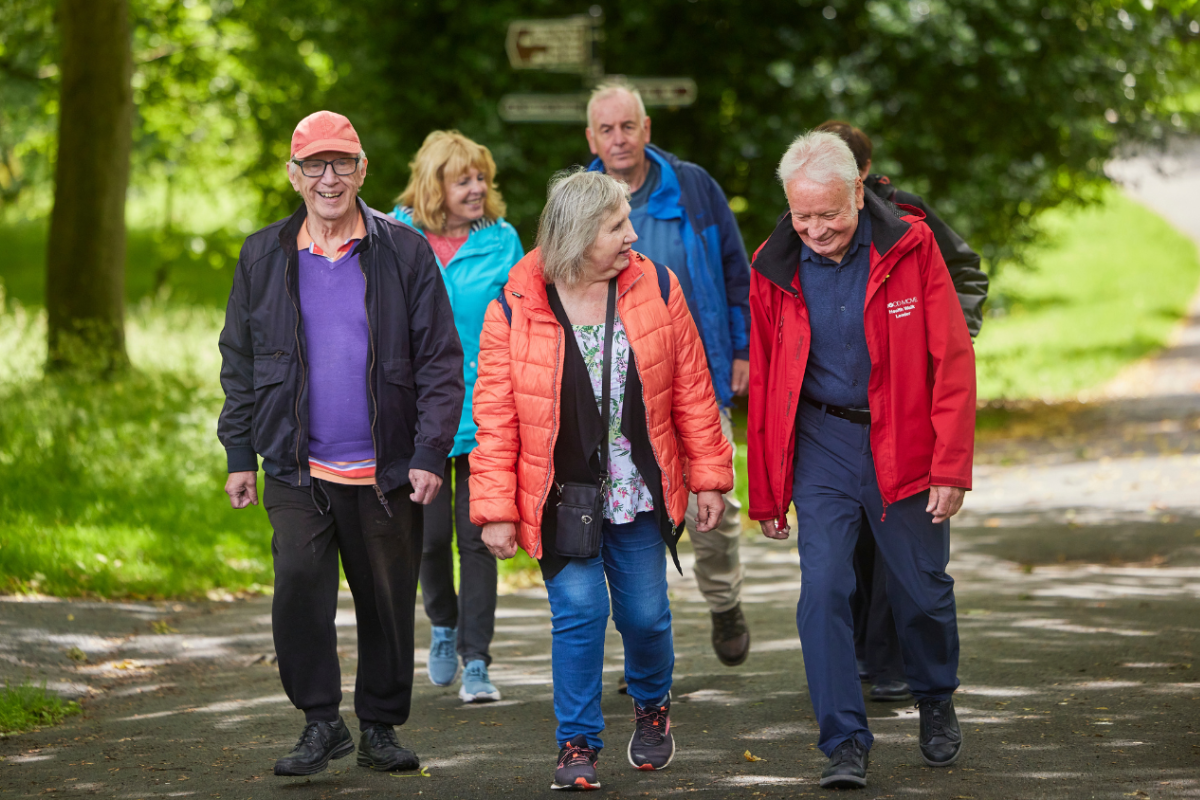 A group of men and woman smile as they walk along a woodland path. Sun shines through the trees behind them.