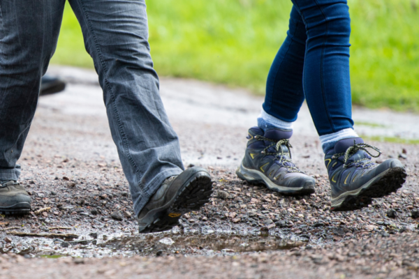 The lower legs and boots of two individuals walking along a gravel path.