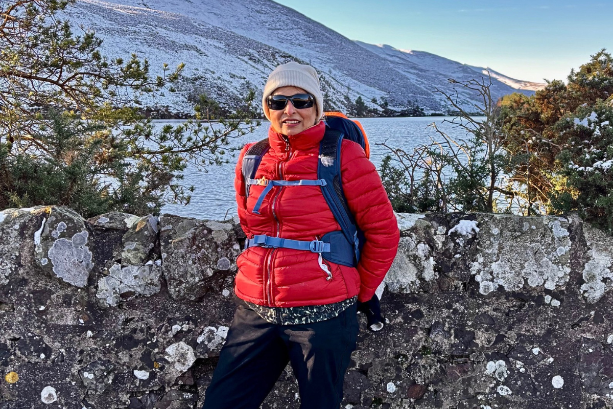 A woman in a red jacket, hat and glasses smiles as she stands near a stone wall, with hillside stretching out into the distance behind,