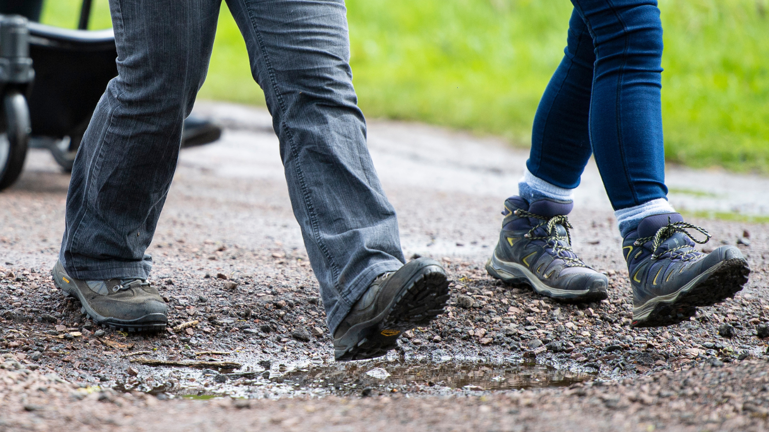 The lower legs and boots of two individuals walking along a gravel path.