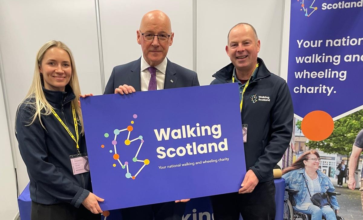 Two staff from Walking Scotland and the First Minister, John Swinney, smile and hold a board with the Walking Scotland logo on it