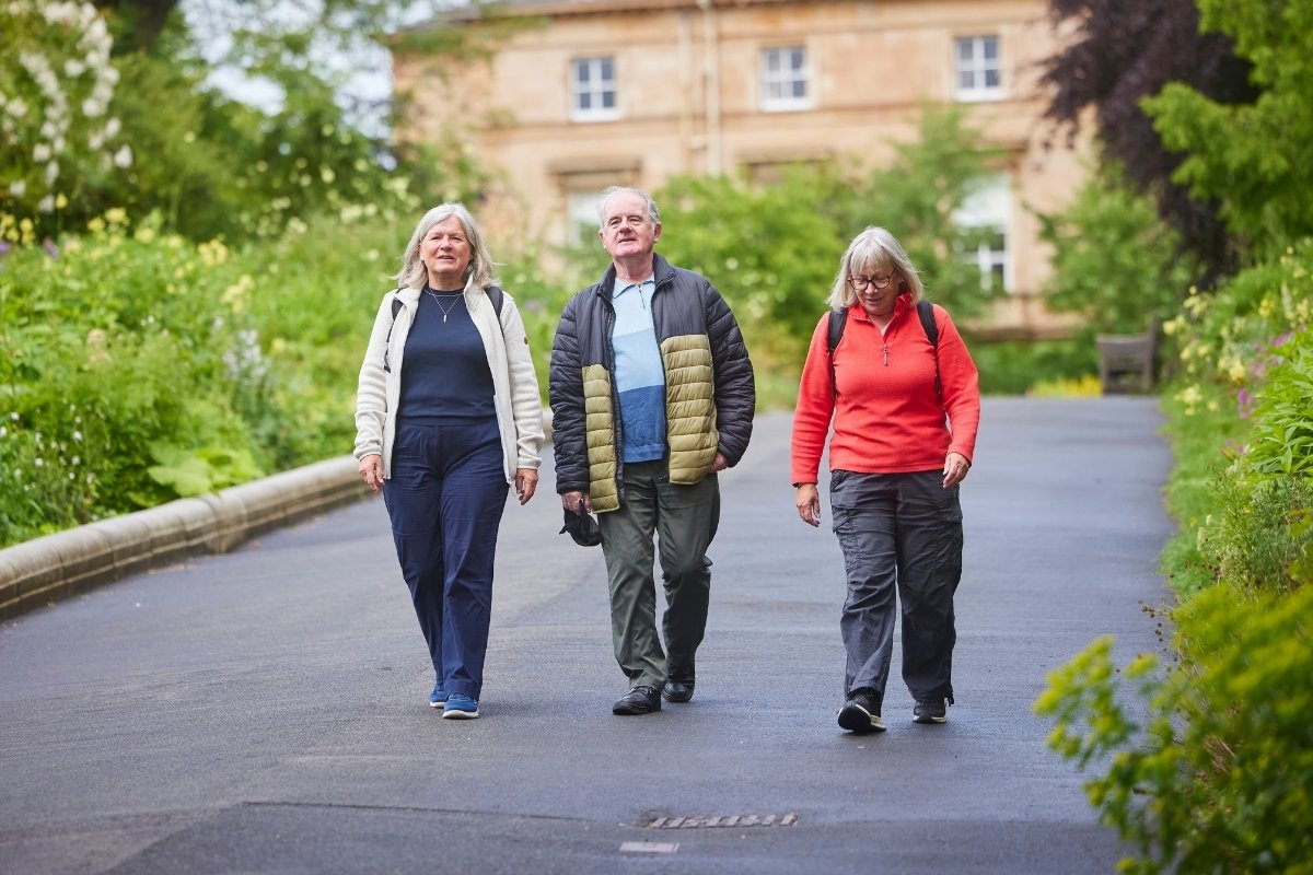 Two older women and an older man smile as they walk along a path between well-tended gardens. A large house can be seen behind them.