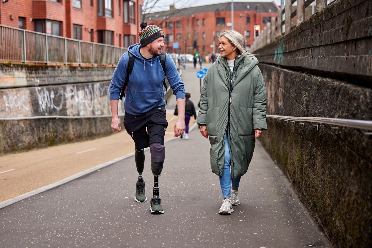 A gentleman with prosthetic legs smiles as he walks alongside a woman with blonde hair and a long beige jacket. They walk along a shared use path in a busy urban area.