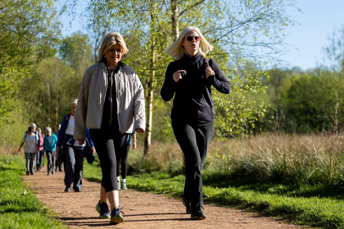 Two women in comfortable walking outfits walk along a path through a local park. They lead a small group of walkers.