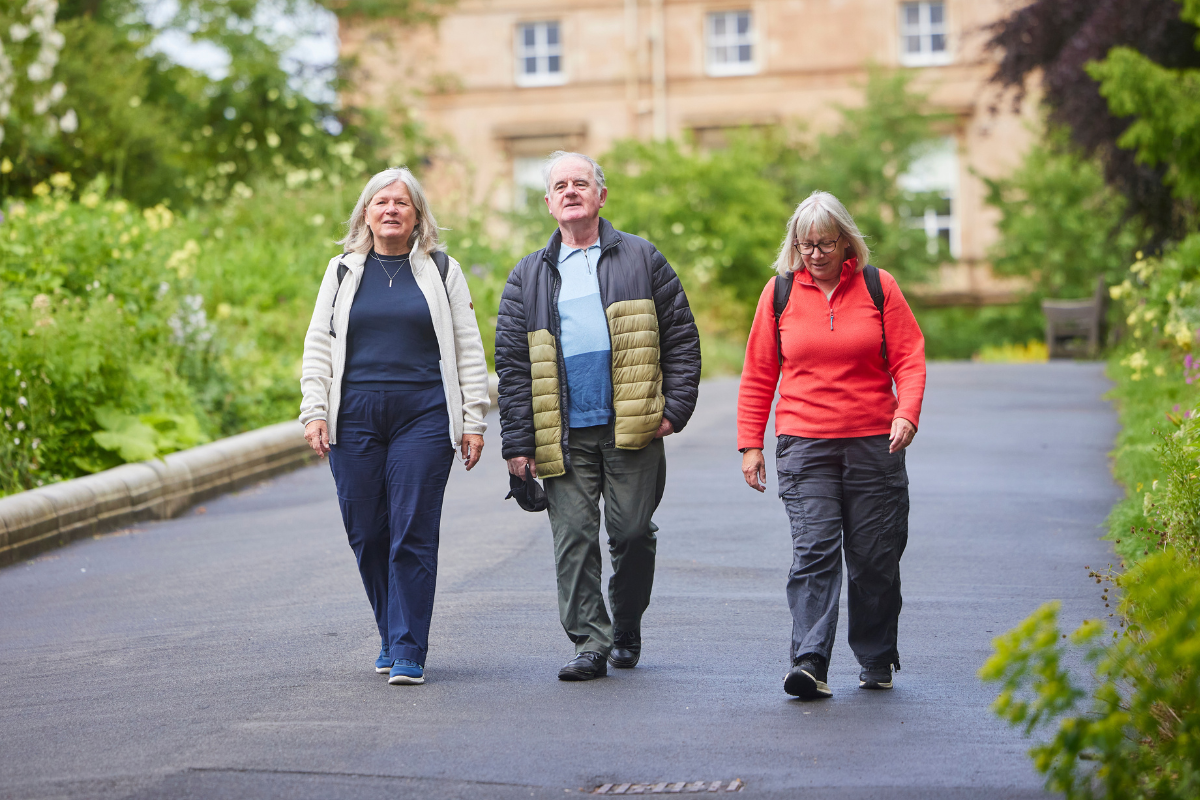 Two older women and an older man smile as they walk along a path between well-tended gardens. A large house can be seen behind them.