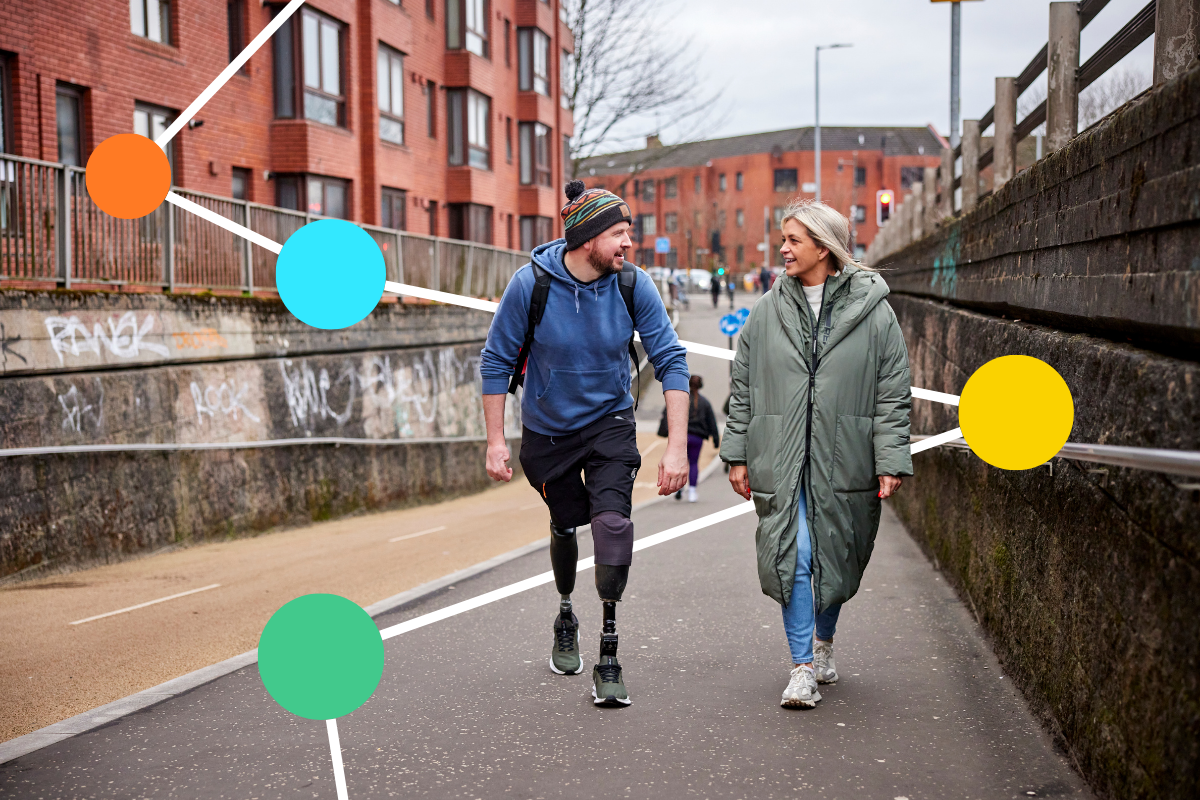 A man with two prosthetic legs smiles as he walks alongside a woman. They walk along a shared path in an urban area.