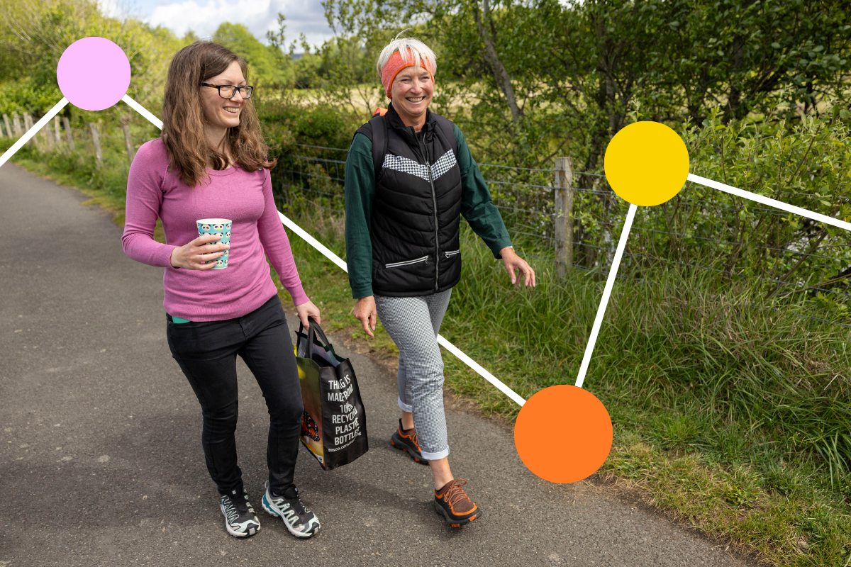 Two women smile as they walk along a path bordered by grass and trees. One woman is carrying a coffee mug and a shopping bag.
