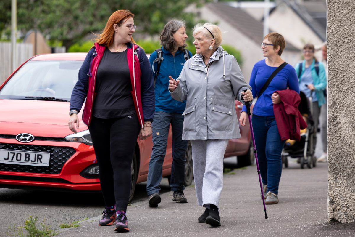 Four women walking in pairs talk as they walk along a street lined with parked cars.