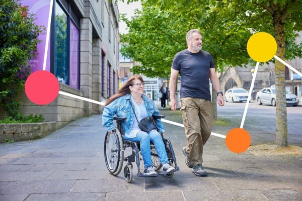 A woman in a wheelchair and a man smile as they walk and wheel side by side along a street.