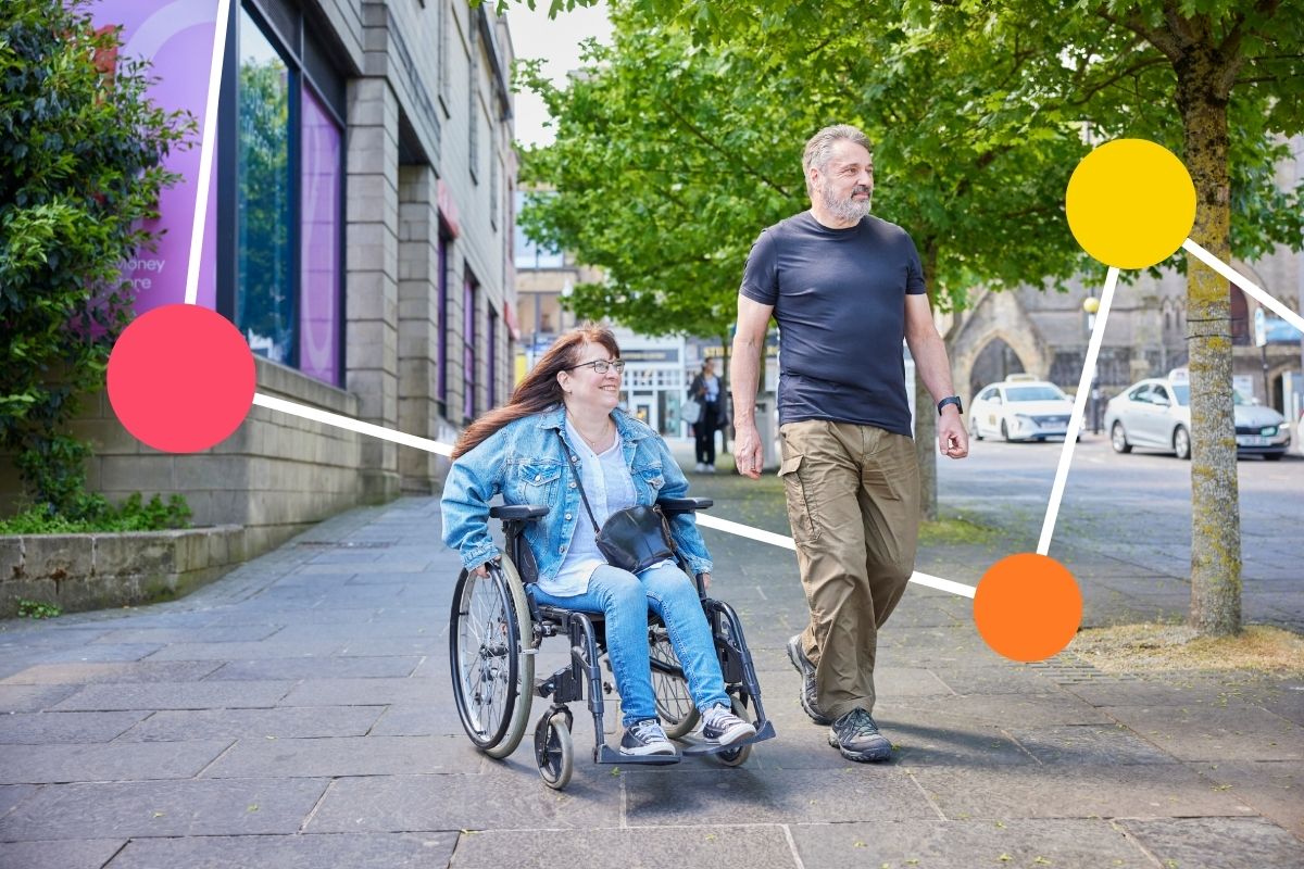A woman in a wheelchair and a man smile as they walk and wheel side by side along a street.