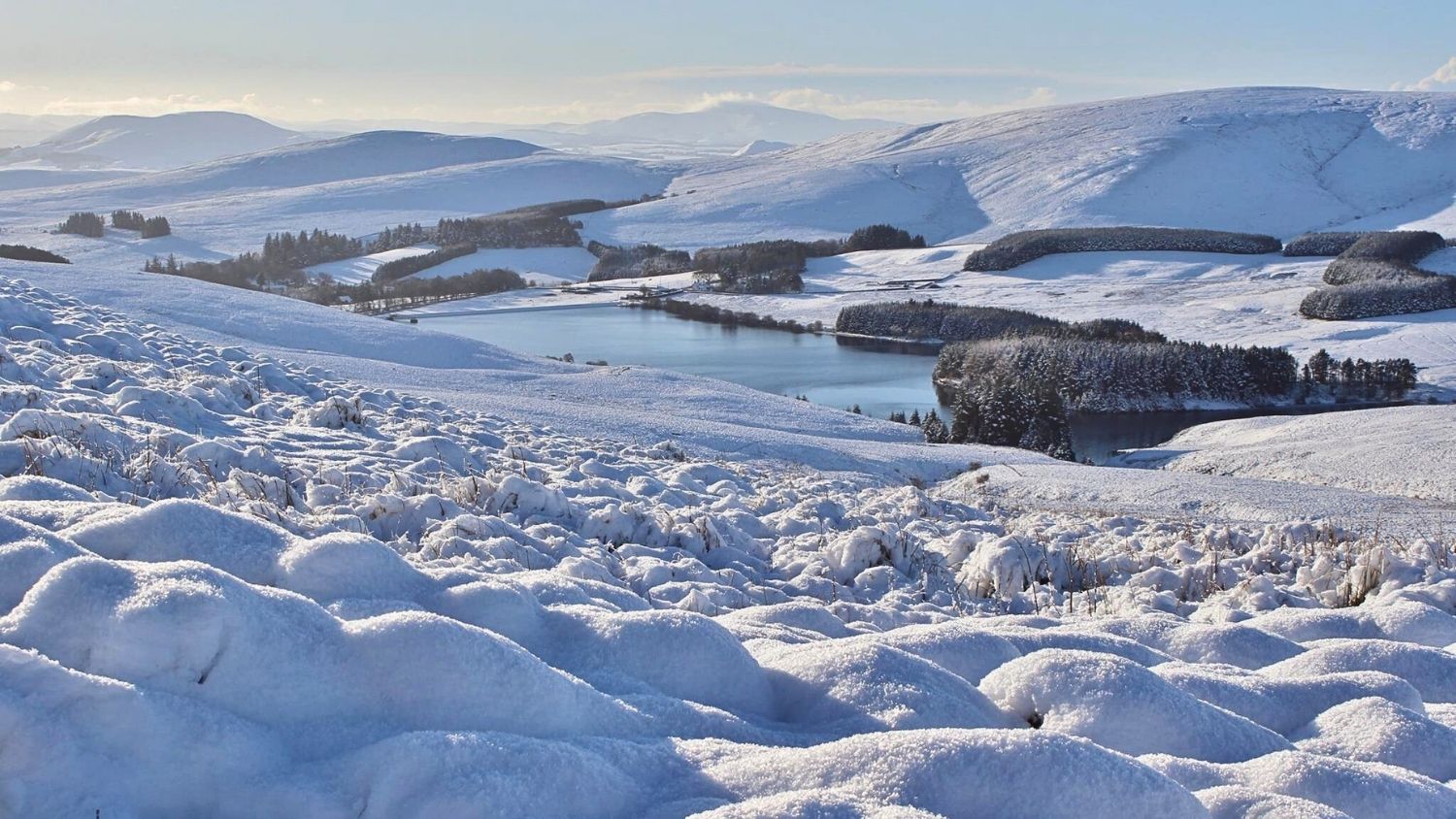 A snow covered valley of rolling hills and woodland.