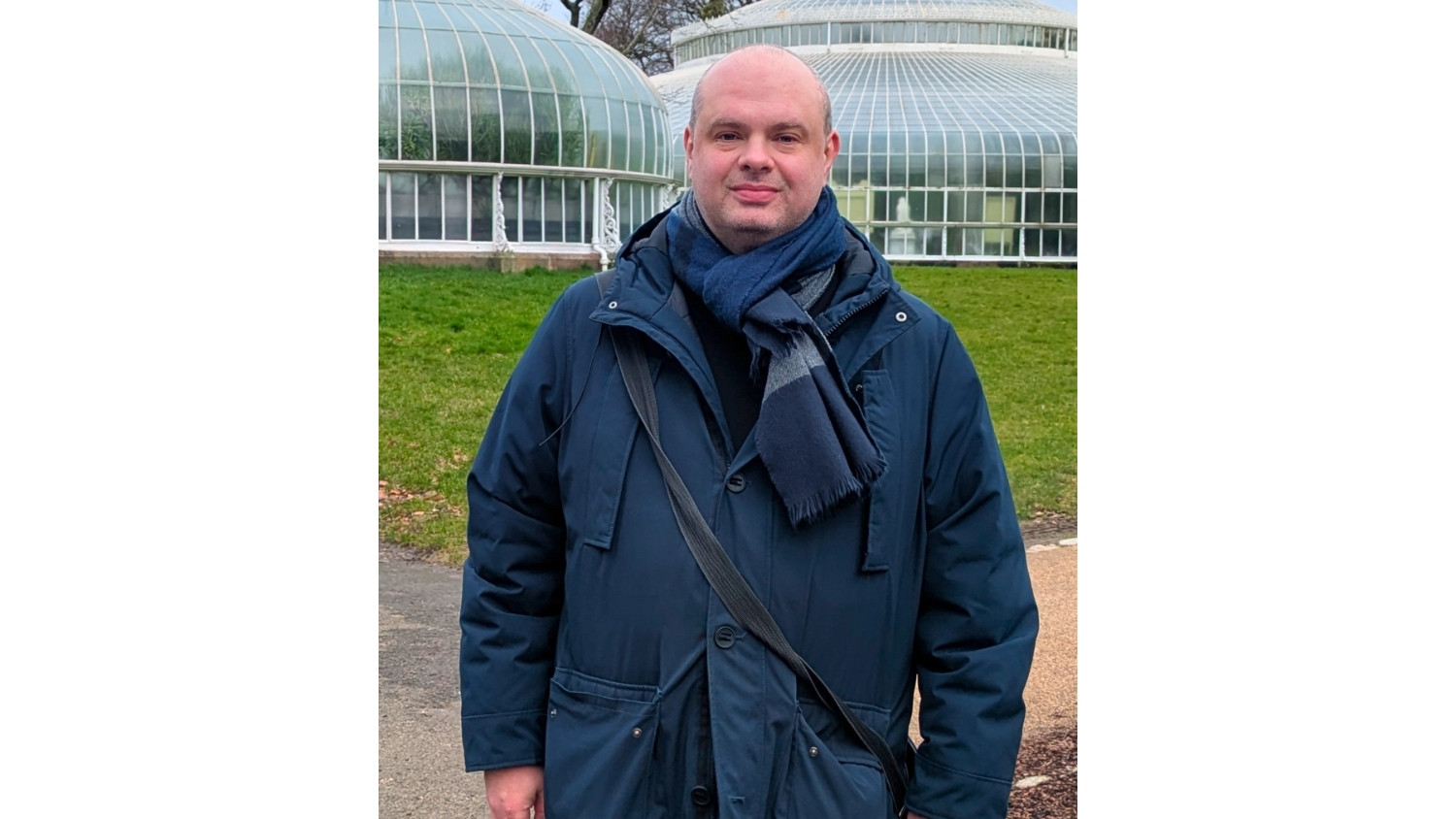 A man smiles at the camera. He's wearing a navy blue coat and scarf. 