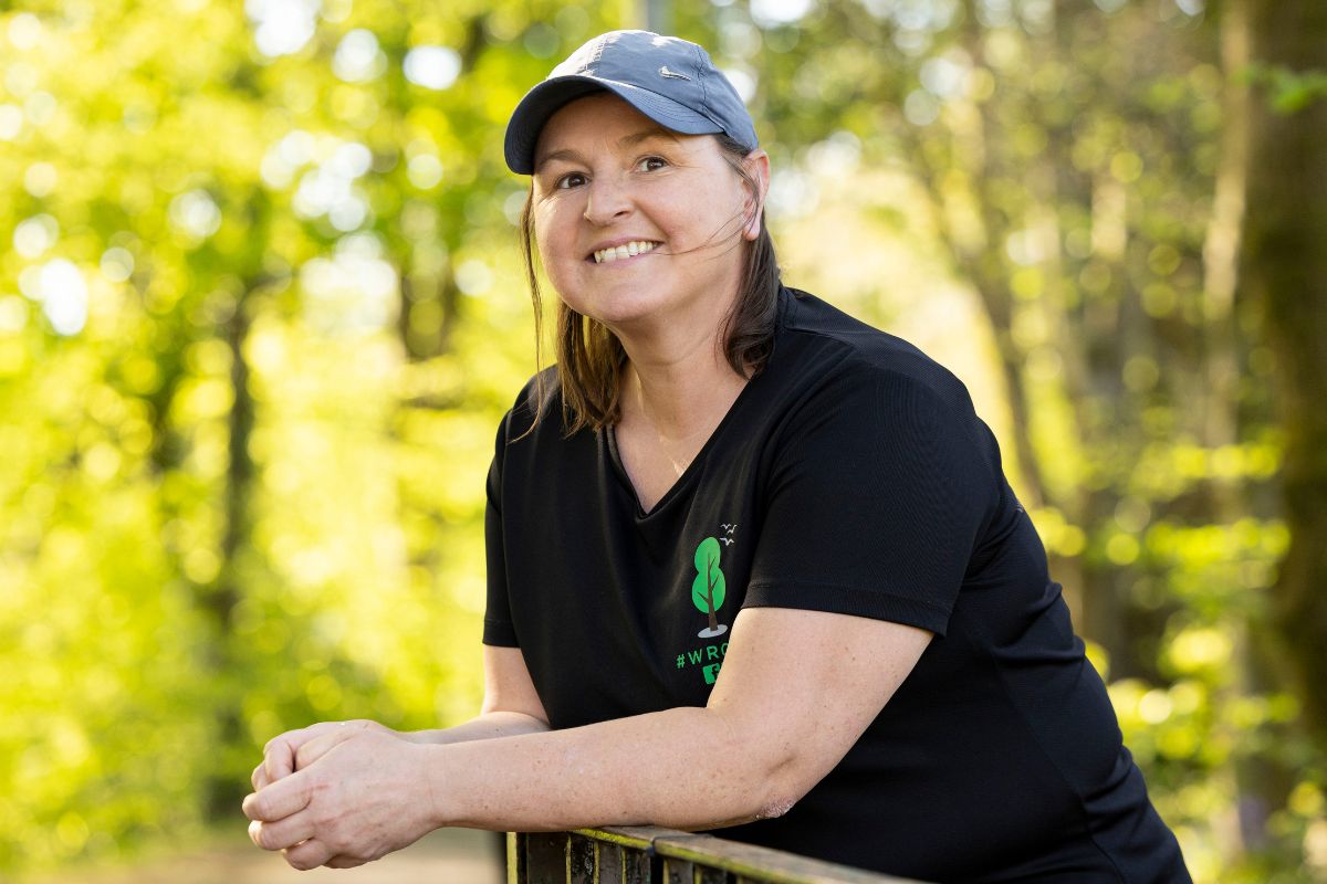 Lorna leans against a railing, smiling at the camera. There are trees in the background.