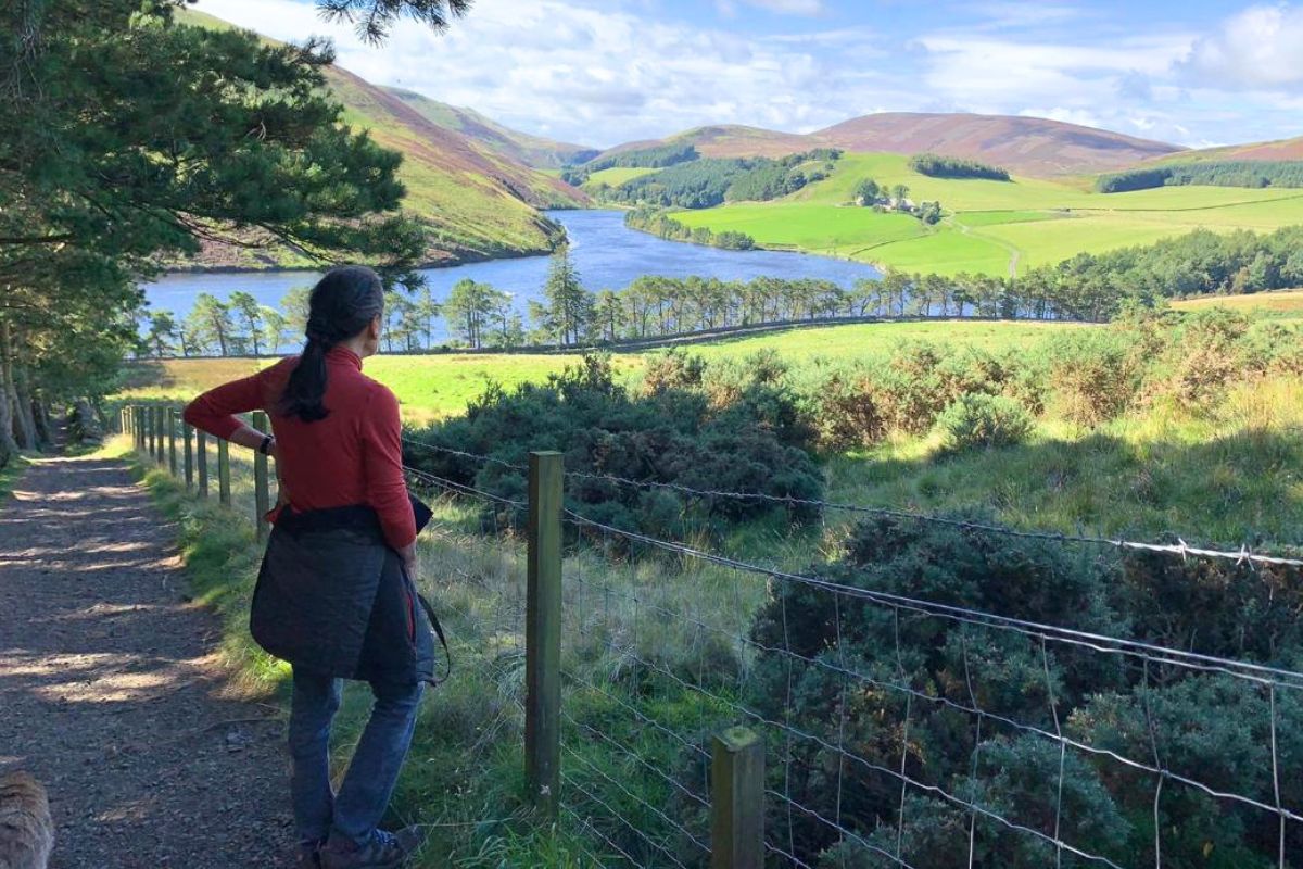 A woman in a red fleece looks out over the view of a loch and rolling hills.