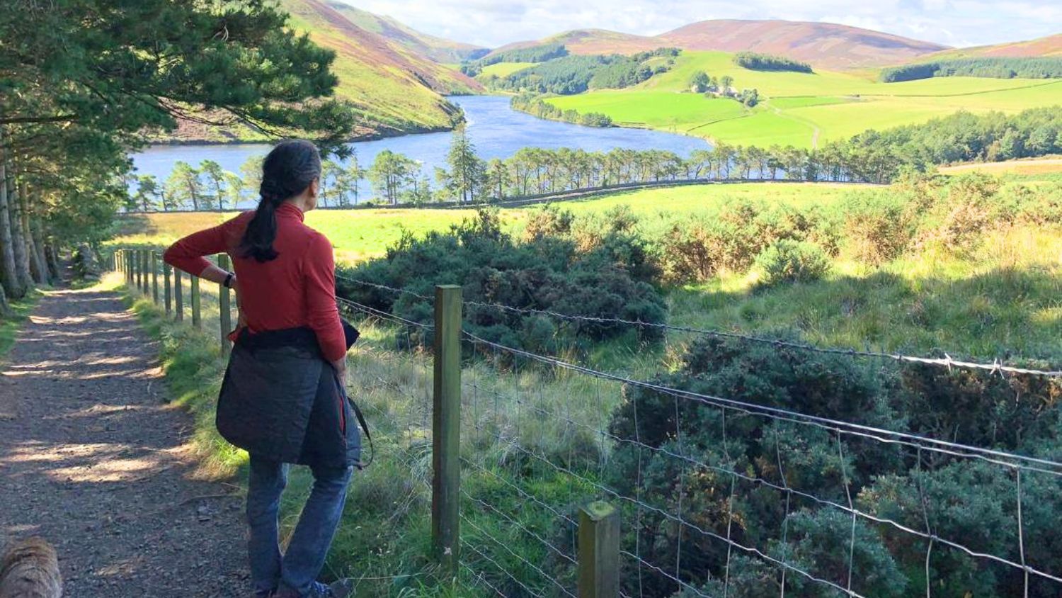 A woman in a red fleece looks out of the view of a loch and rolling hills. 