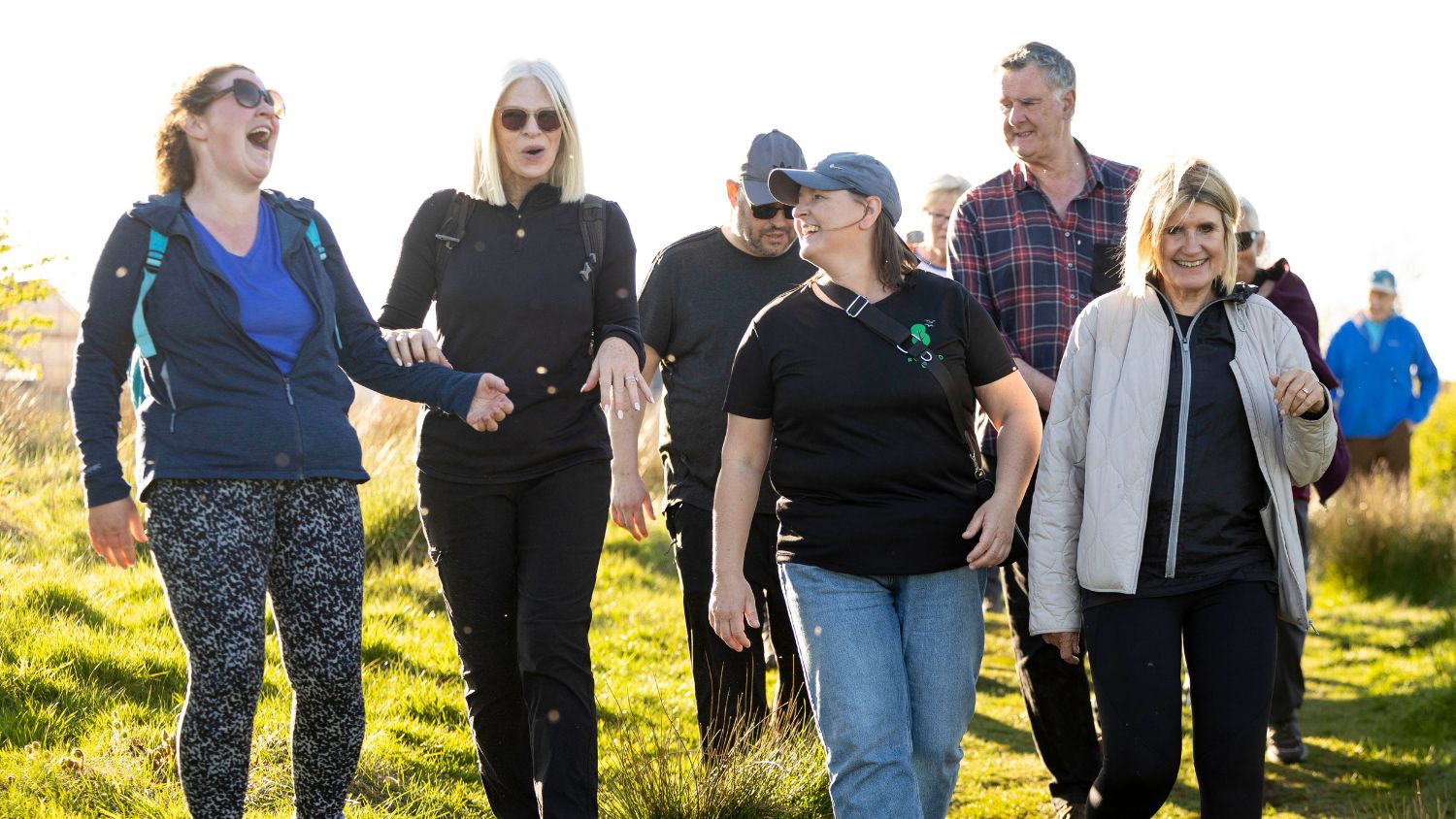 A group of people walk through a field. They are laughing together. They're wearing walking clothing. 