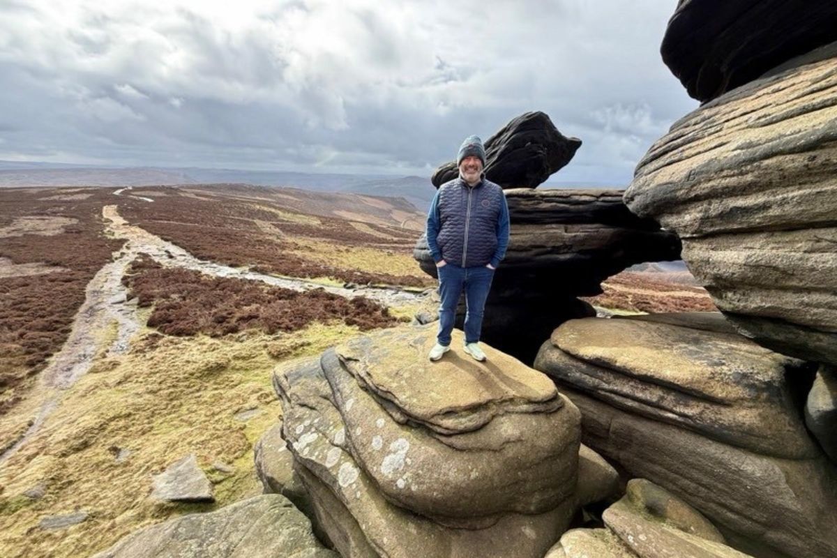 A man stands smiling on top of a rock. The landscape is the background shows hills and shrubbery.