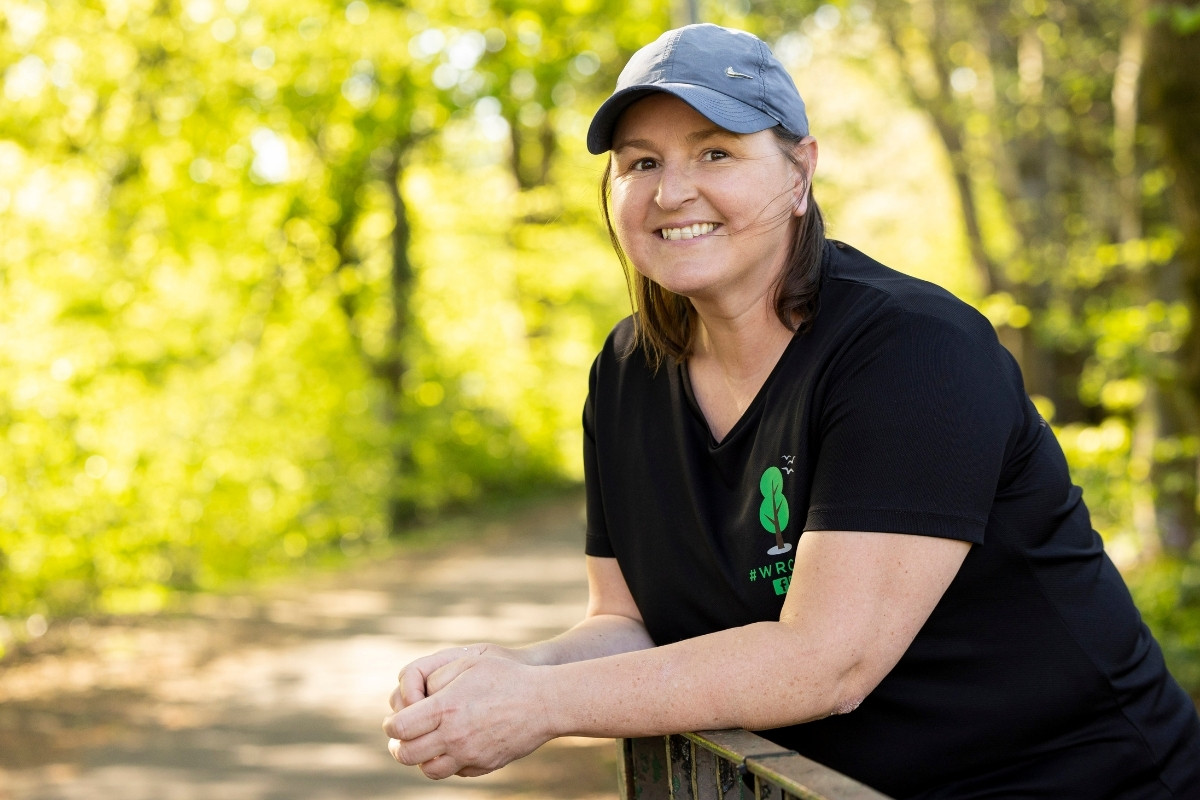 Lorna, a woman wearing a black t-shirt and grey cap, smiles as she leans on a railing.