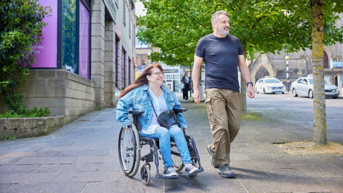 A woman in a wheelchair and a man smile as they walk and wheel side by side along a street.