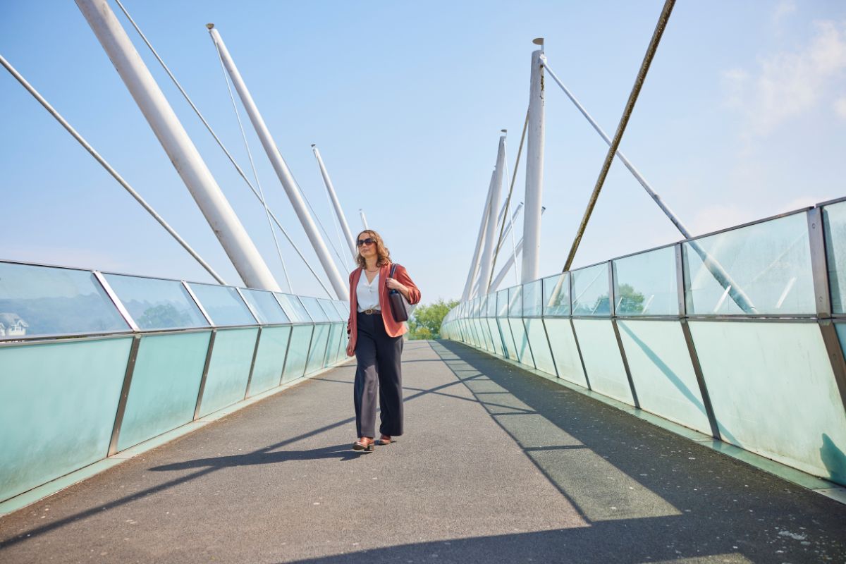 A woman walks along a bridge. She is wearing sunglasses and a red jacket.