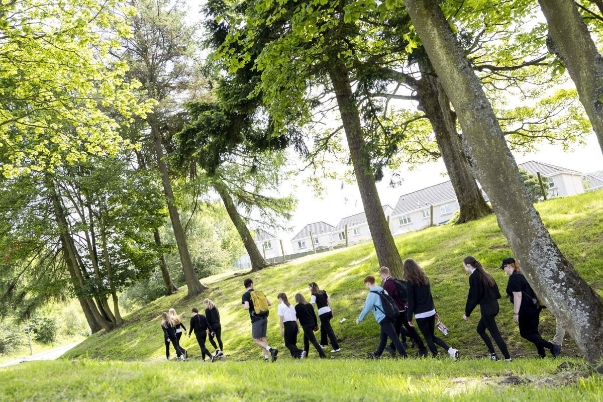 A group of young people in school uniform walk along a path. There is greenery and fields.