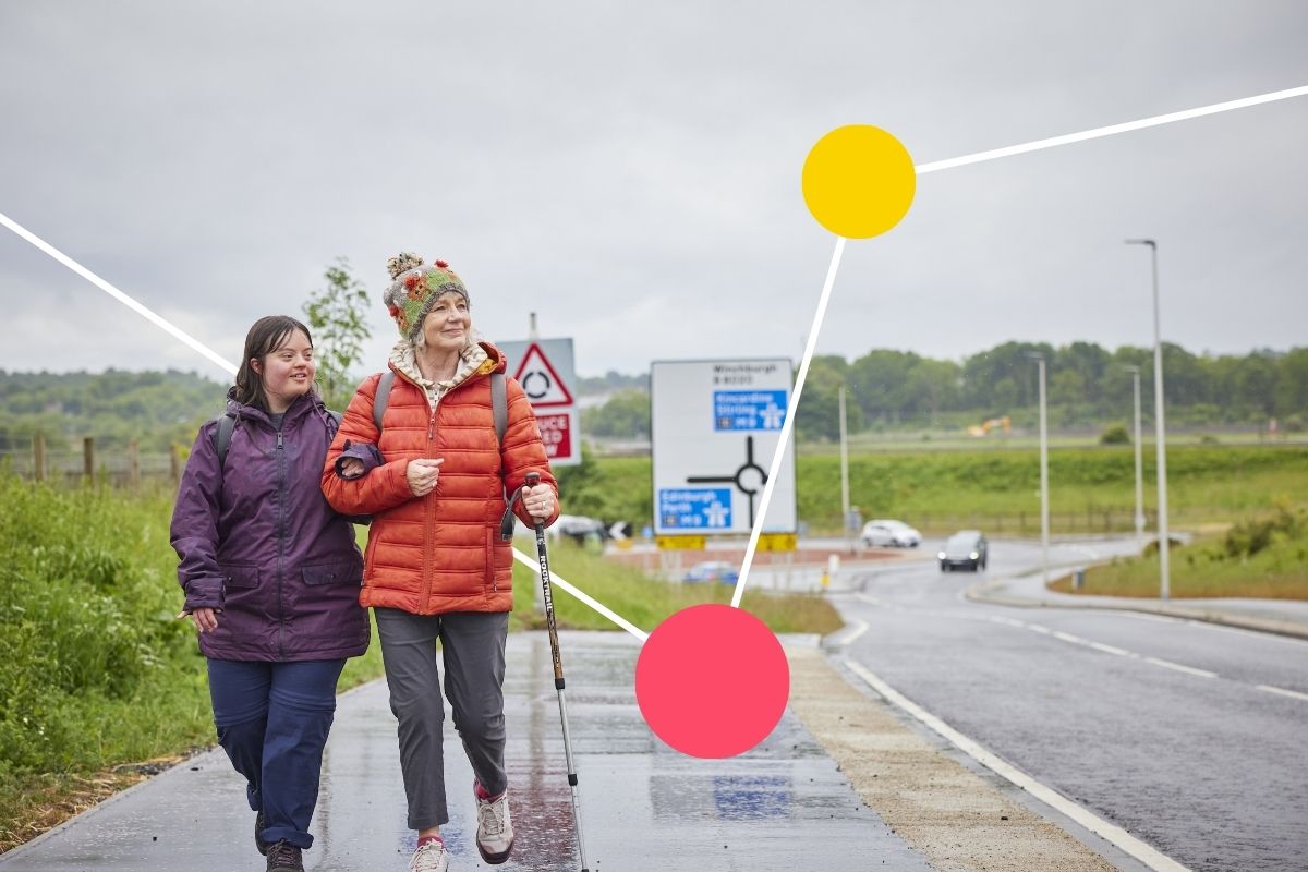 2 women walk arm in arm along a wide path by the side of the road. It is raining lightly and they are wearing waterproof coats.