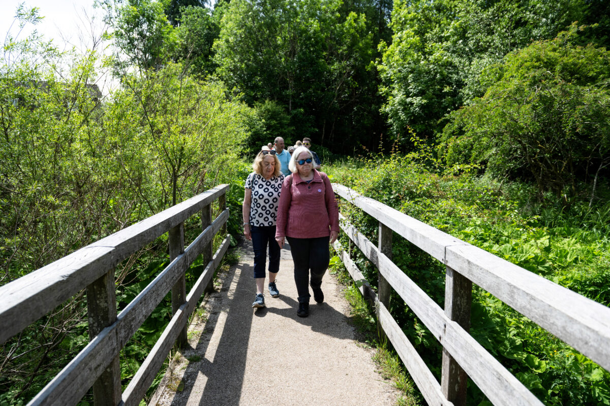 2 women cross a wooden bridge. There is greenery either side.