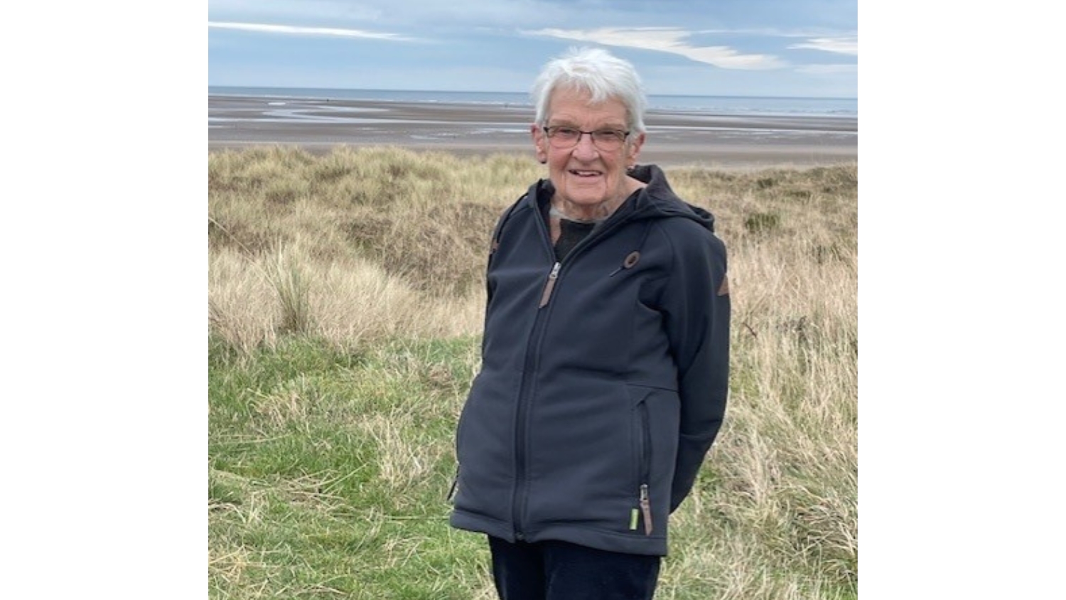 Rae stands on a beach, smiling at the camera. She has short white hair and is wearing a black jacket and black trousers. 
