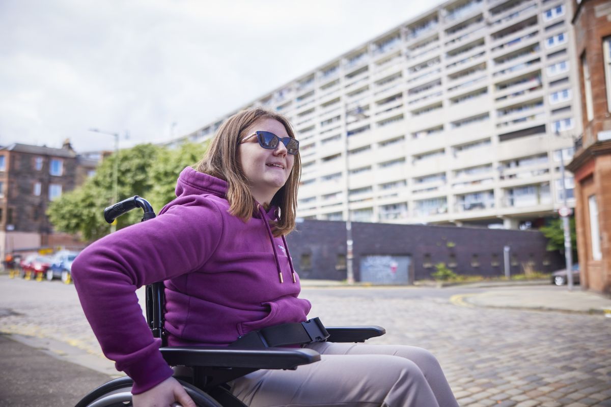A woman wearing a purple hoodie and sunglasses smiles. She is using a wheelchair along a city street.