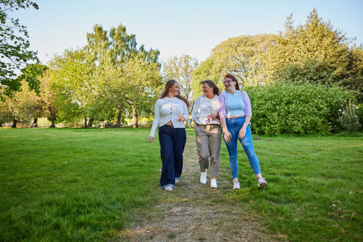 3 women walk in a park on a sunny day. They are chatting and laughing.