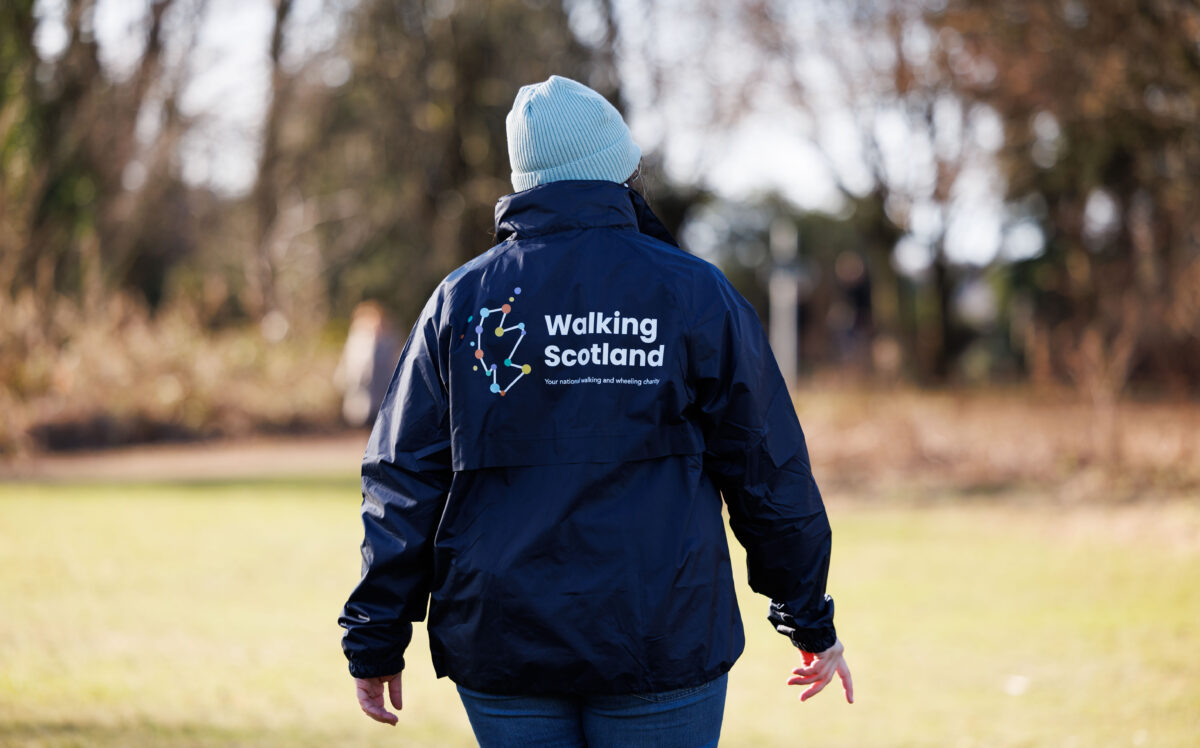 Image is of a man walking in field, his back is to the camera. The logo of Walking Scotland on his rain jacket is the centre focus.